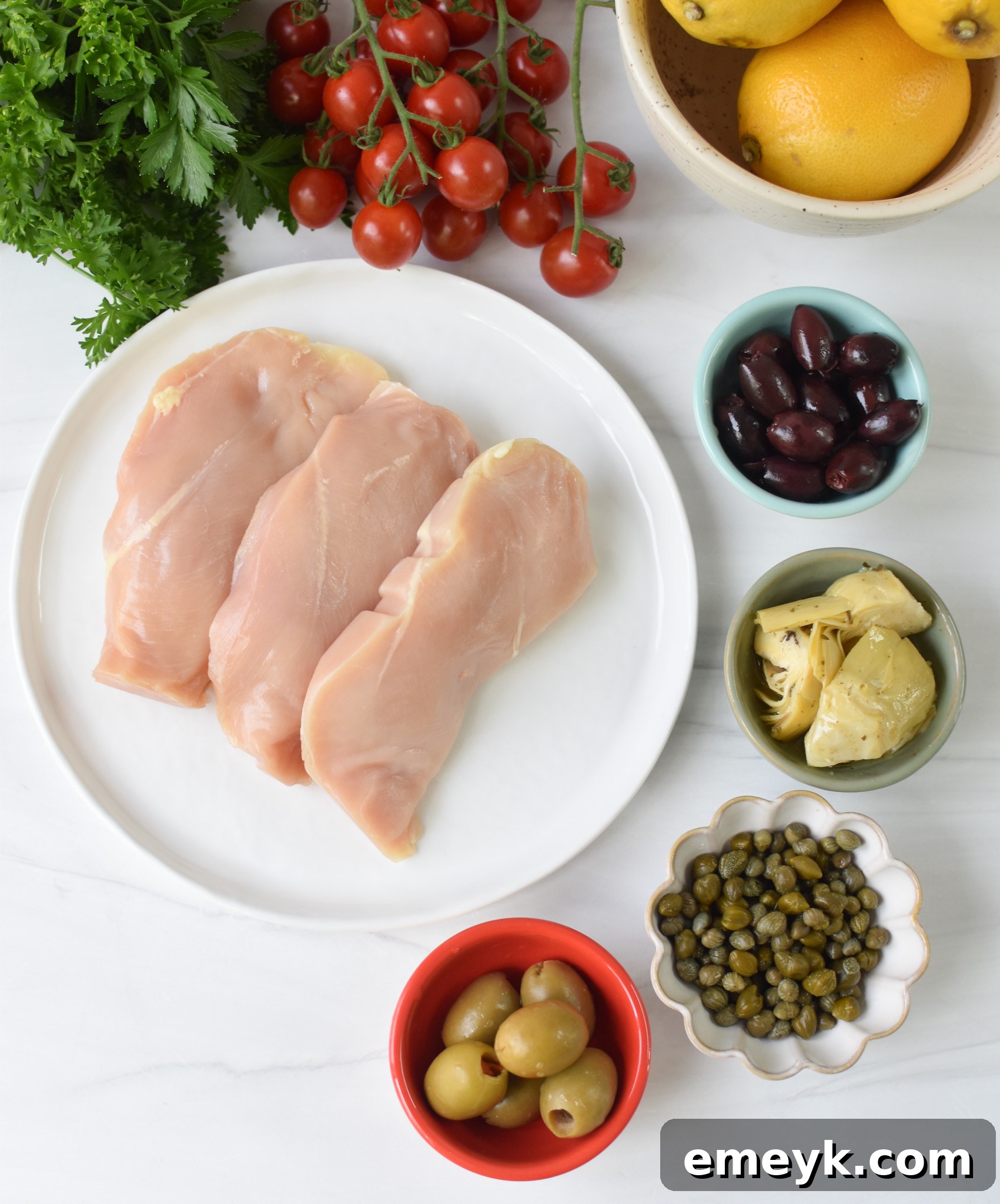 A collection of fresh ingredients laid out for Mediterranean Skillet Chicken, including chicken breasts, lemon, herbs, olives, and tomatoes.