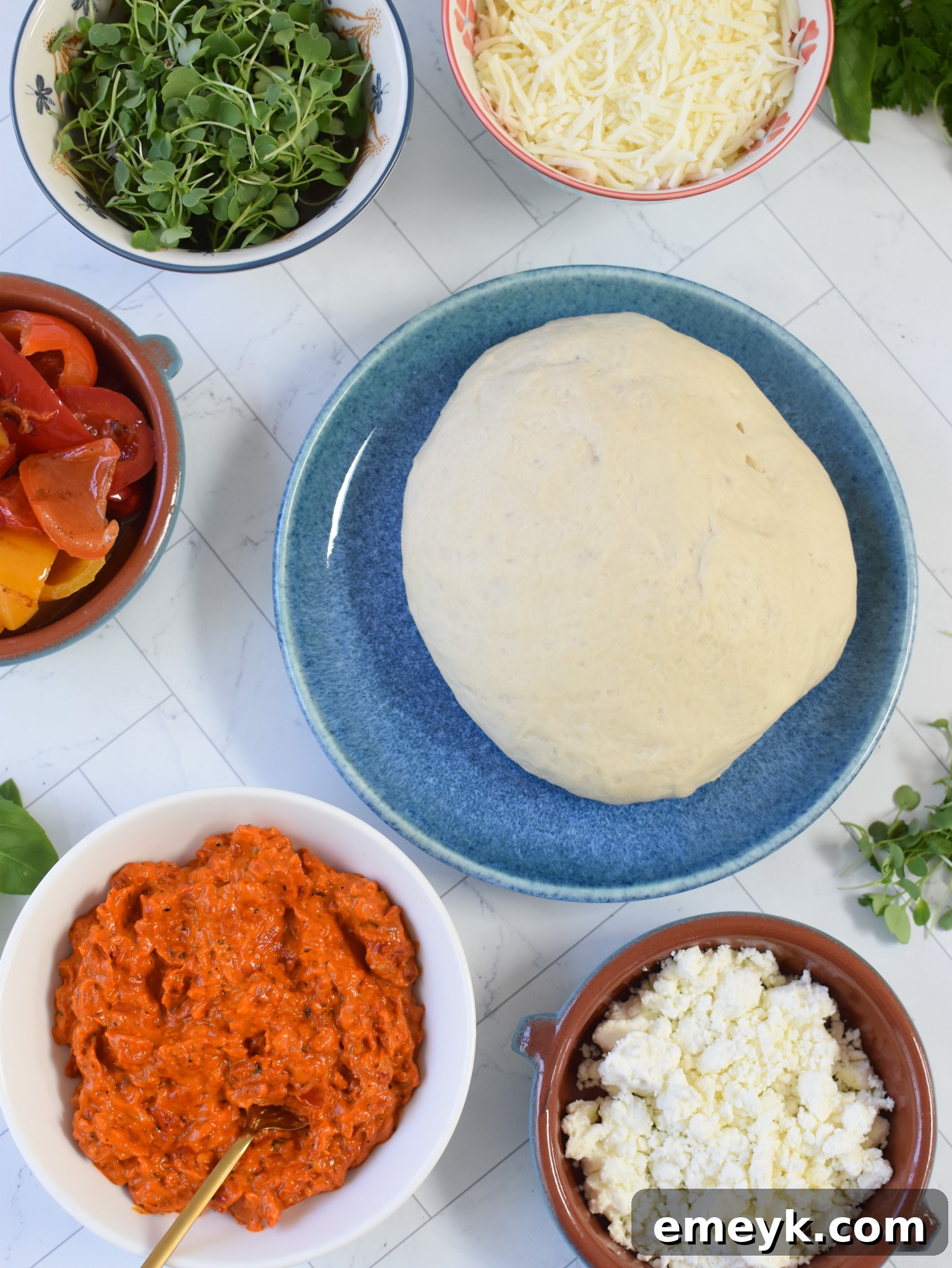 A vibrant flat lay of all the fresh ingredients required for Cast Iron Mediterranean Pizza, including dough, a jar of red pepper spread, various cheeses, and roasted red peppers.