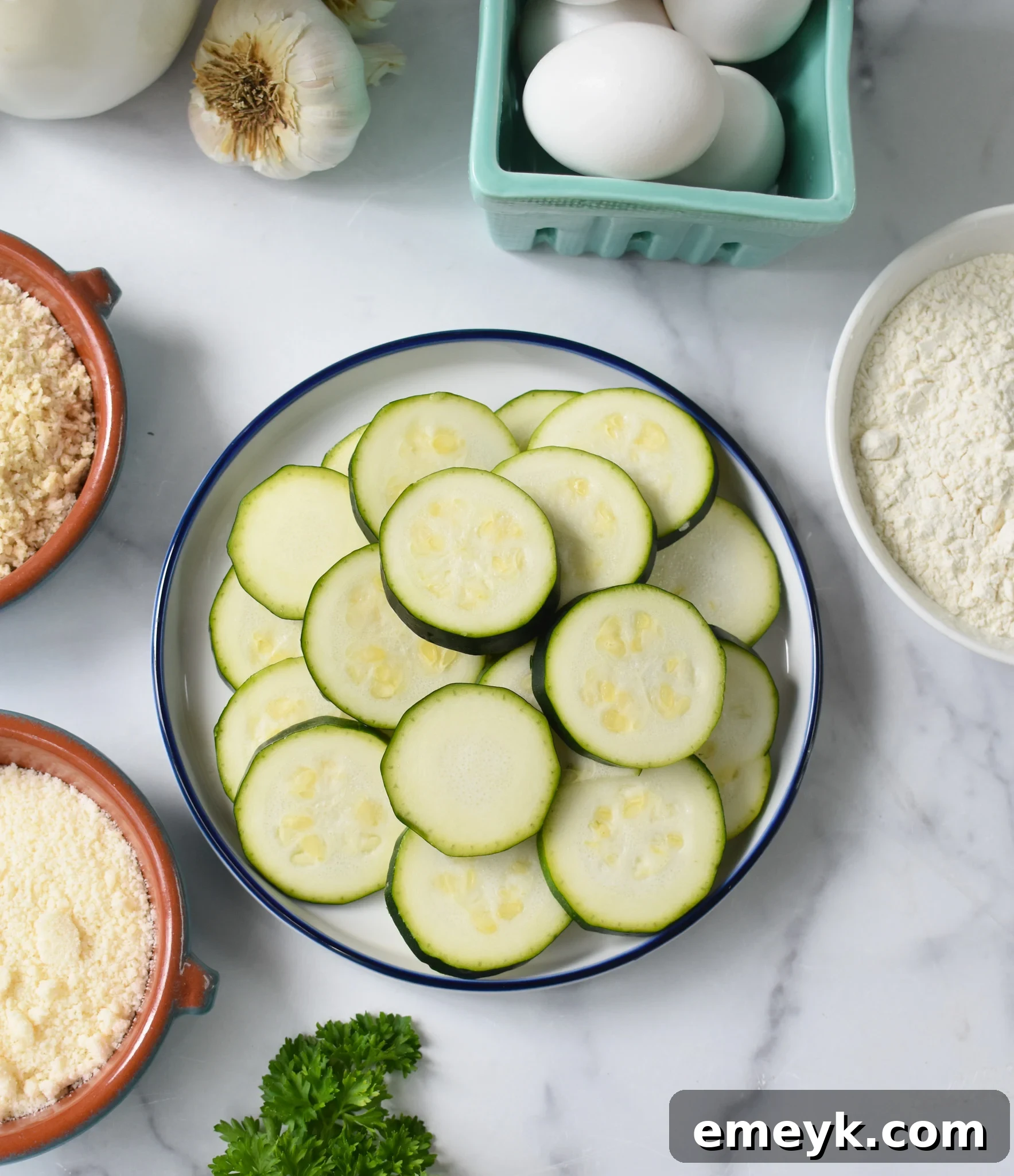 A collection of fresh ingredients laid out for Air Fryer Zucchini Chips, including zucchini, eggs, flour, breadcrumbs, and spices.