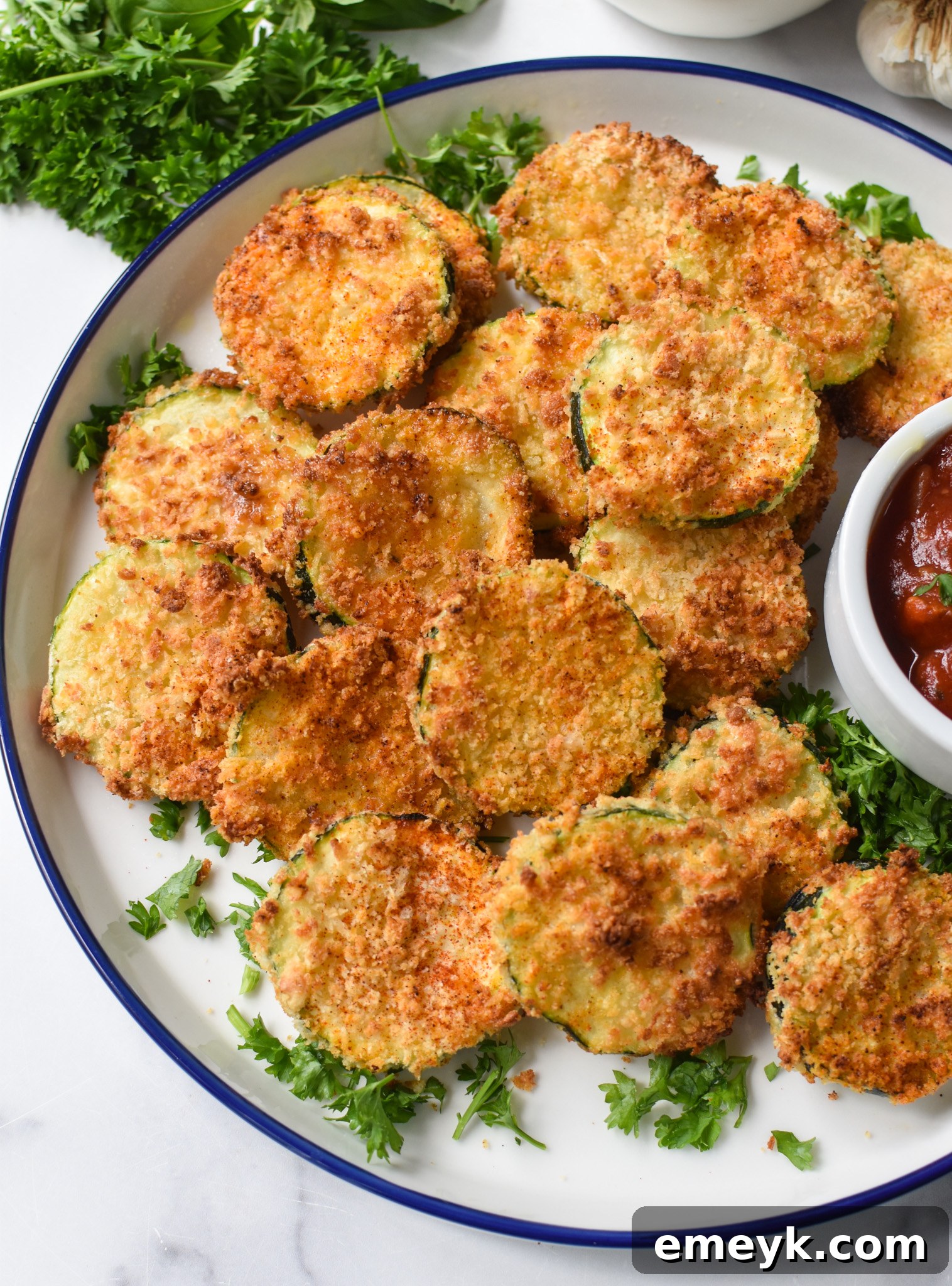 A vibrant plate of Air Fryer Zucchini Chips garnished with fresh parsley, alongside a small bowl of marinara sauce for dipping.
