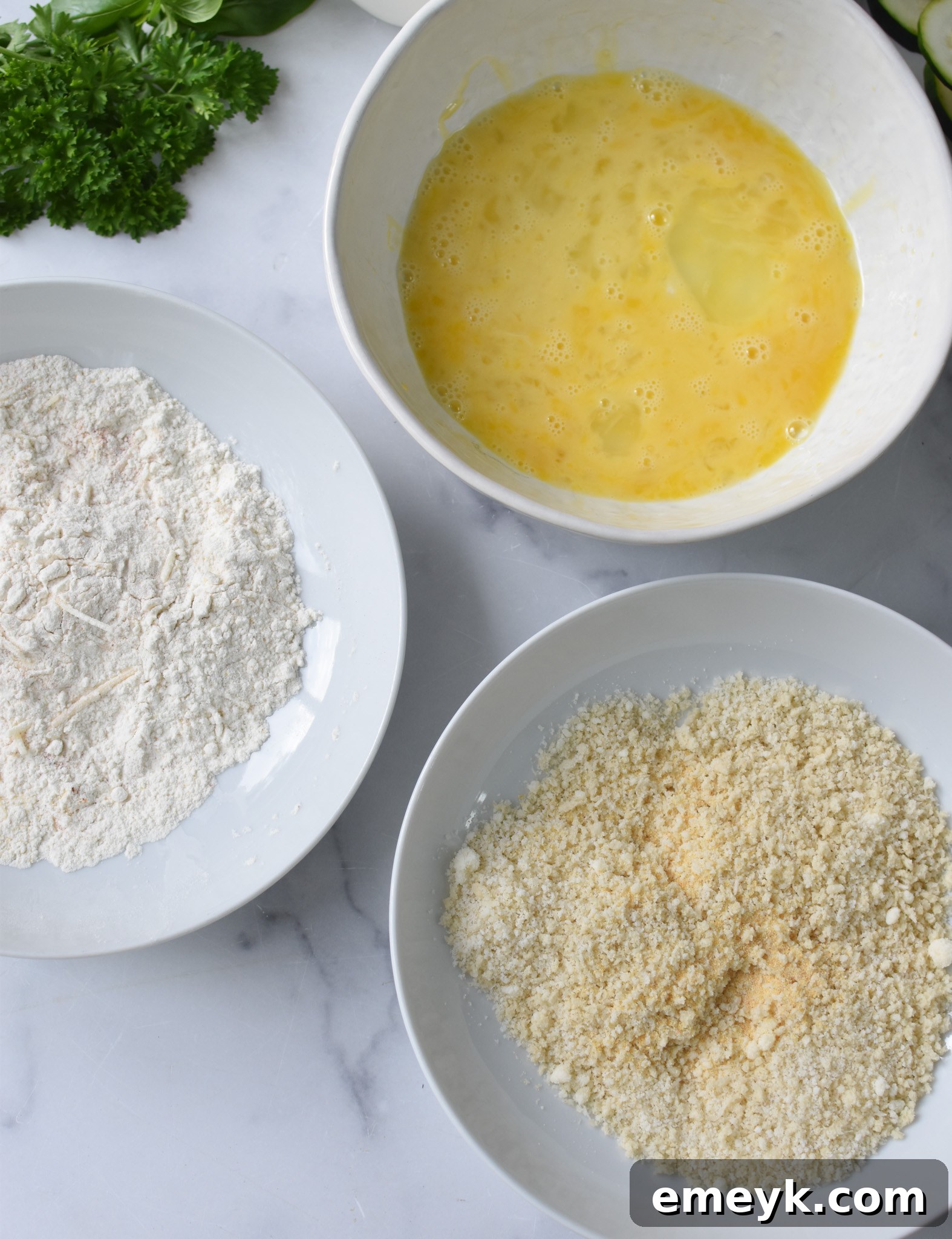 Three separate bowls containing the breading ingredients: flour mixture, beaten egg, and panko-Parmesan mixture, set up for coating zucchini.
