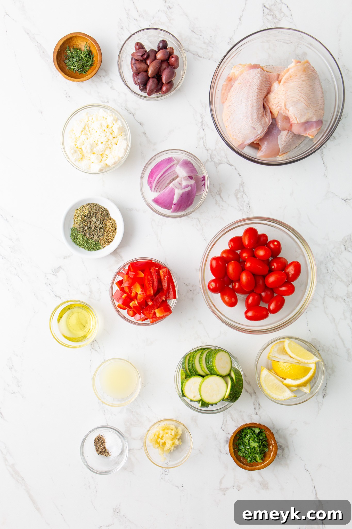 Mediterranean Sheet Pan Chicken ingredients laid out on a table: chicken thighs, red onion, bell pepper, zucchini, feta, olives, cherry tomatoes, and marinade spices.