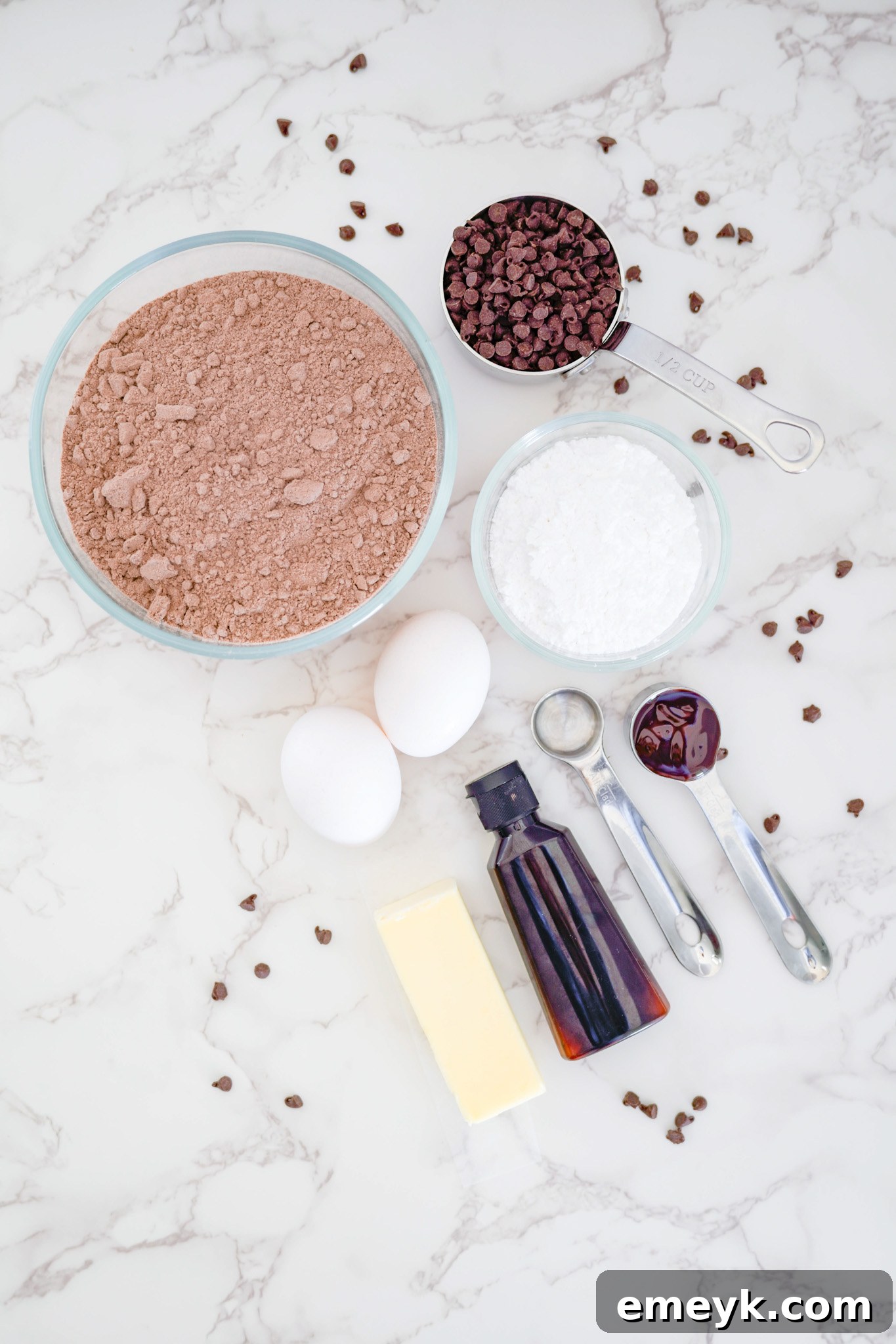 All the necessary ingredients for Chocolate Crinkle Cookies laid out on a table: chocolate cake mix, butter, eggs, vanilla, chocolate syrup, miniature chocolate chips, and powdered sugar.