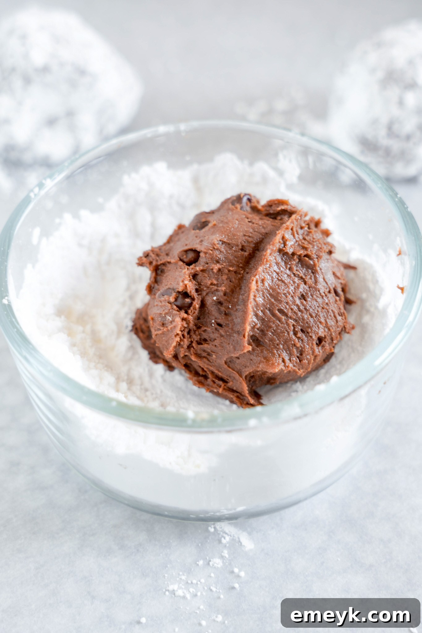 Chocolate Crinkle Cookie dough ball being rolled and coated in powdered sugar in a bowl.