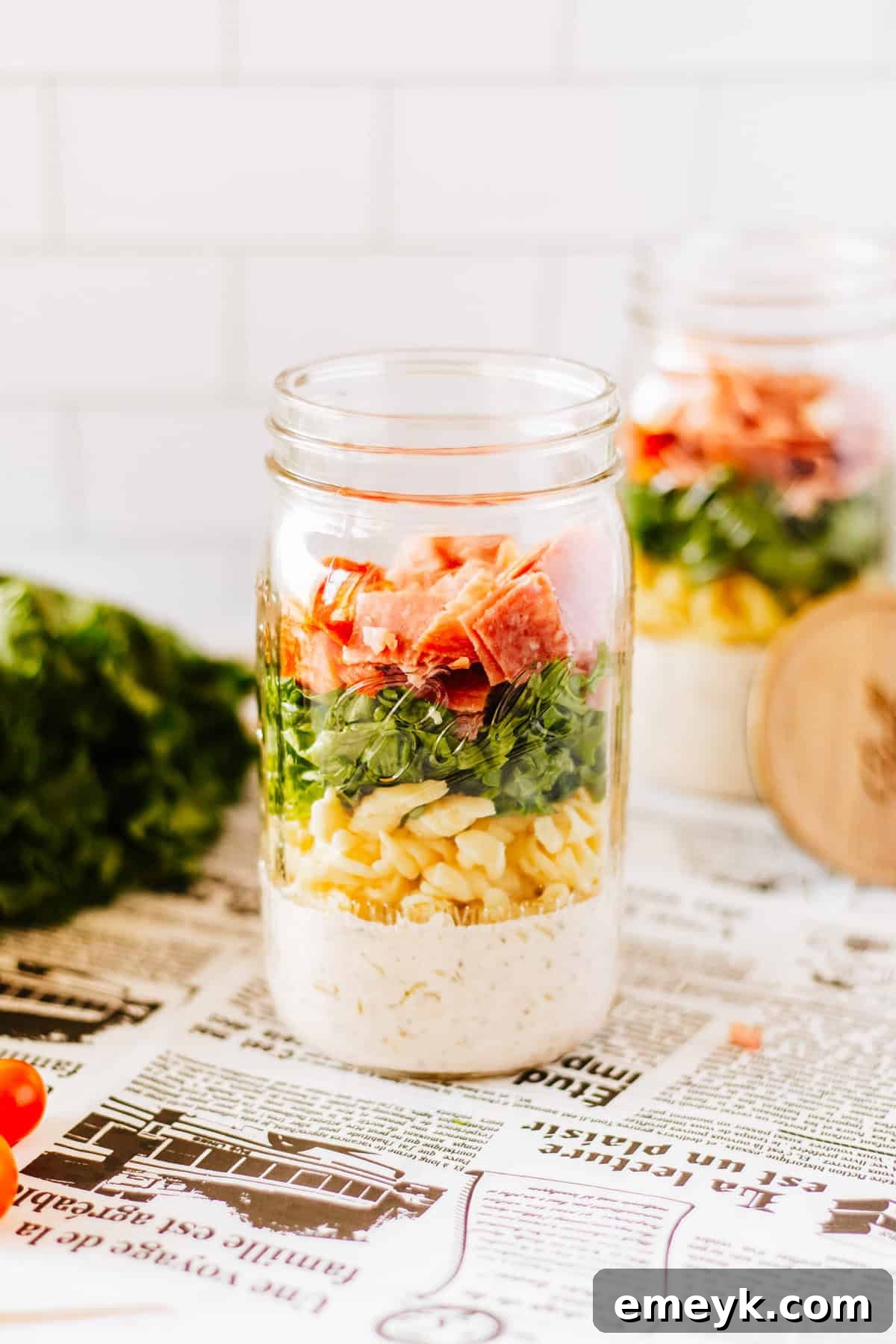 A beautifully layered Italian Pasta Mason Jar Salad in a glass jar, showing distinct sections of dressing, vegetables, pasta, meats, cheese, and lettuce.