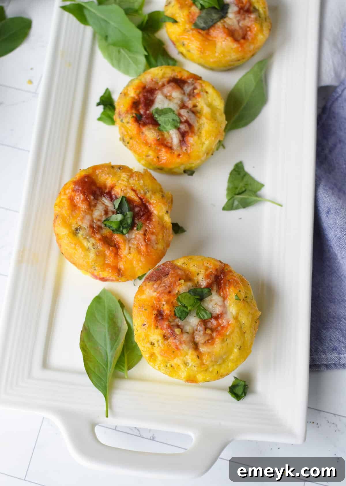 Three golden-brown Pizza Egg Muffins arranged on a pristine white platter, garnished with fresh basil