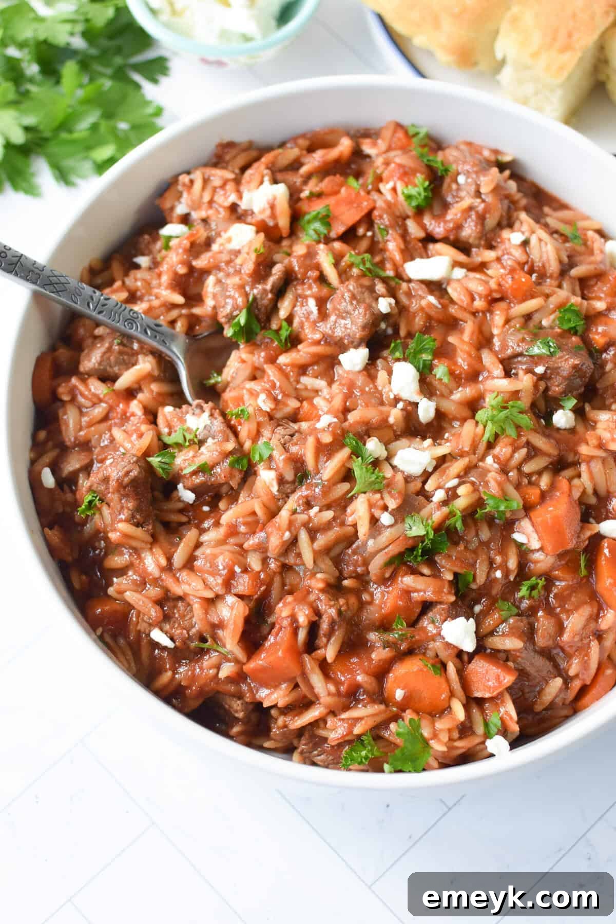 A close-up of a serving of Greek Beef Stew with Orzo in a white bowl, garnished with fresh herbs and crumbled feta cheese.