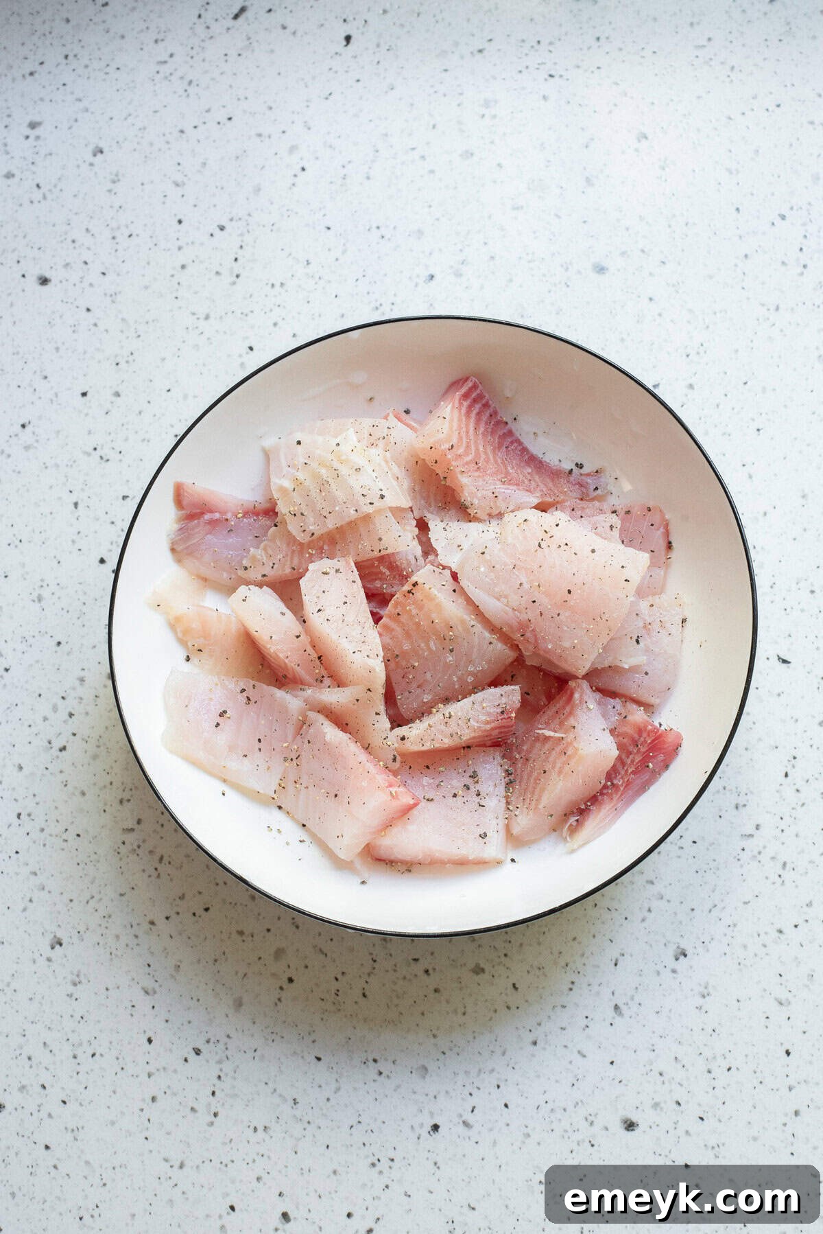 Raw white fish fillets cut into pieces in a white bowl, seasoned with salt and pepper