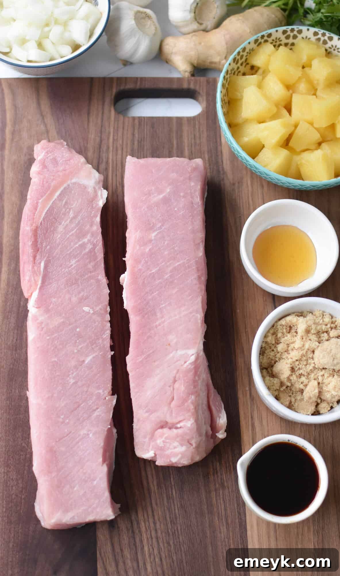 A collection of fresh ingredients laid out, including a pork loin, pineapple chunks, ginger, garlic, onion, and various sauces, ready for the Slow Cooker Pineapple Pork Loin recipe.