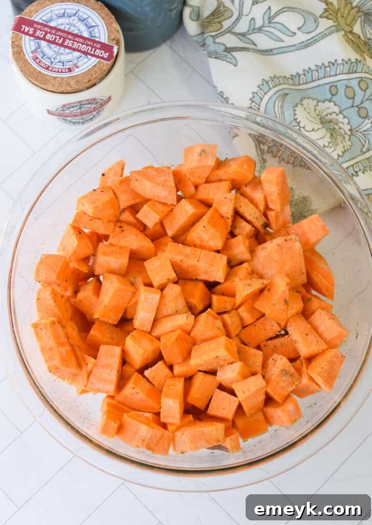 Diced sweet potato chunks tossed with olive oil, salt, and pepper in a clear glass bowl, preparing for roasting.