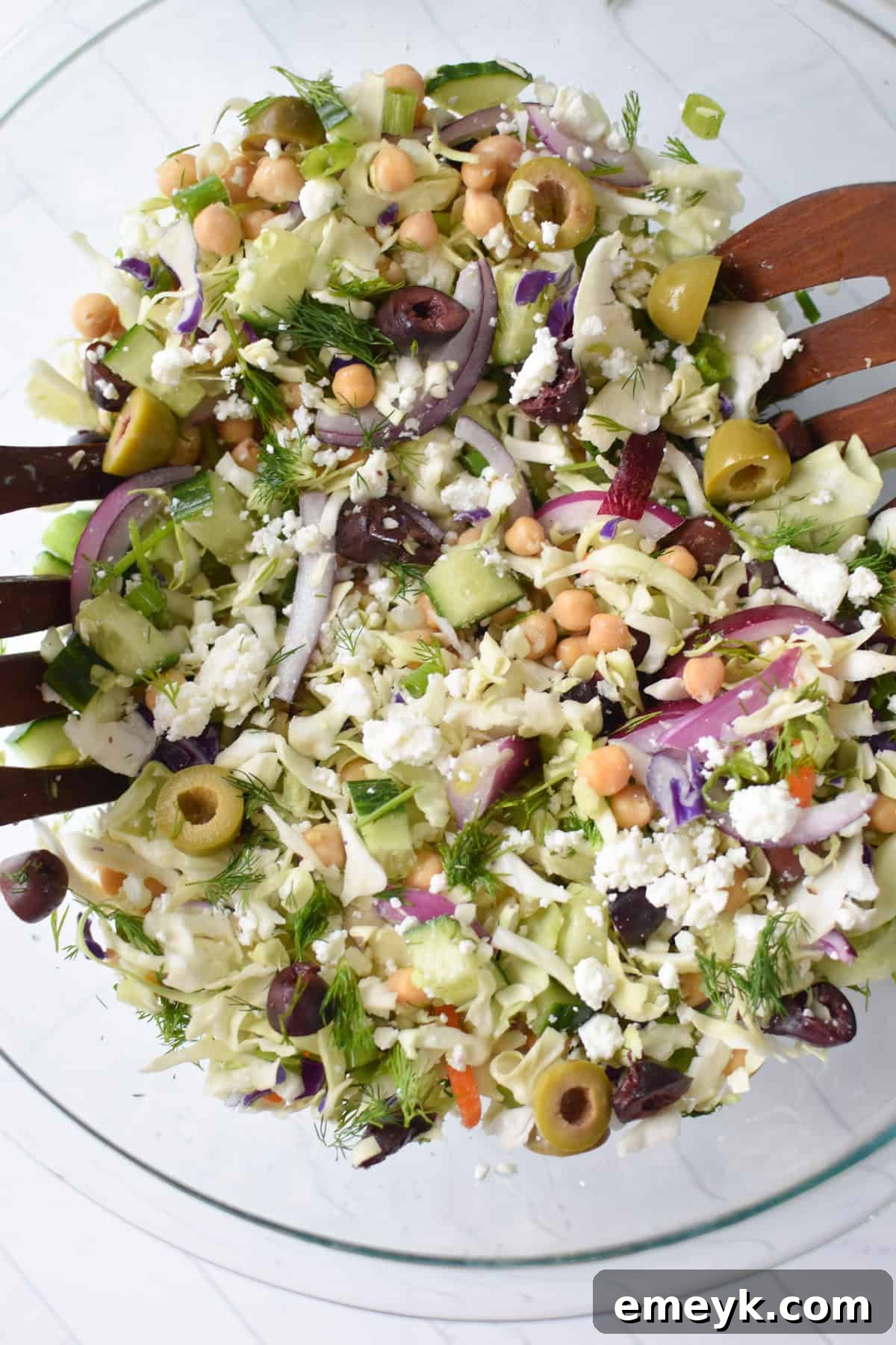 Close-up of Greek Cabbage Salad in a clear glass bowl with serving spoons, highlighting the fresh ingredients and vibrant colors.