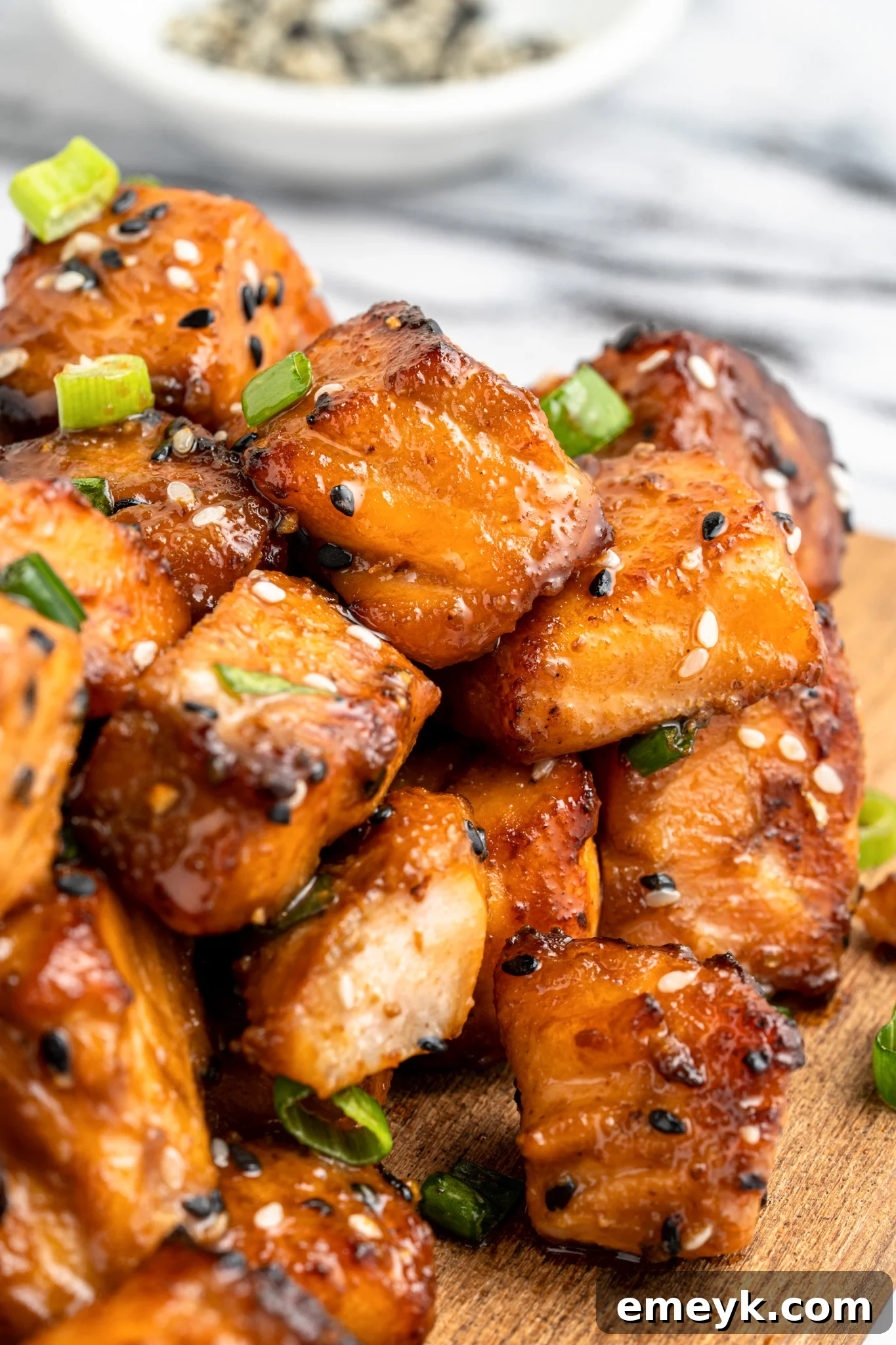 Close-up of glazed salmon cubes garnished with sesame seeds and green onions on a wooden board. A small bowl of sesame seeds is blurred in the background.