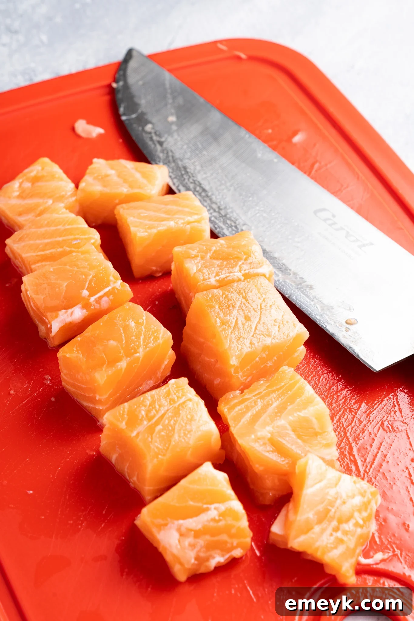 Cubes of fresh salmon are neatly arranged on a bright red cutting board. A large kitchen knife with a shiny blade is positioned next to the salmon pieces. The background has a textured, cool-toned surface.