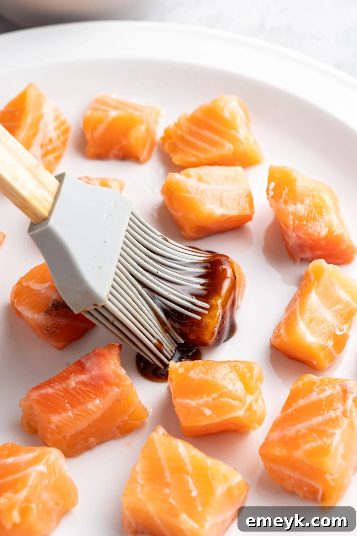 A silicone brush applies a dark marinade to raw salmon cubes on a white plate. The salmon pieces are evenly spaced and glistening, suggesting preparation for cooking. The background is softly blurred.