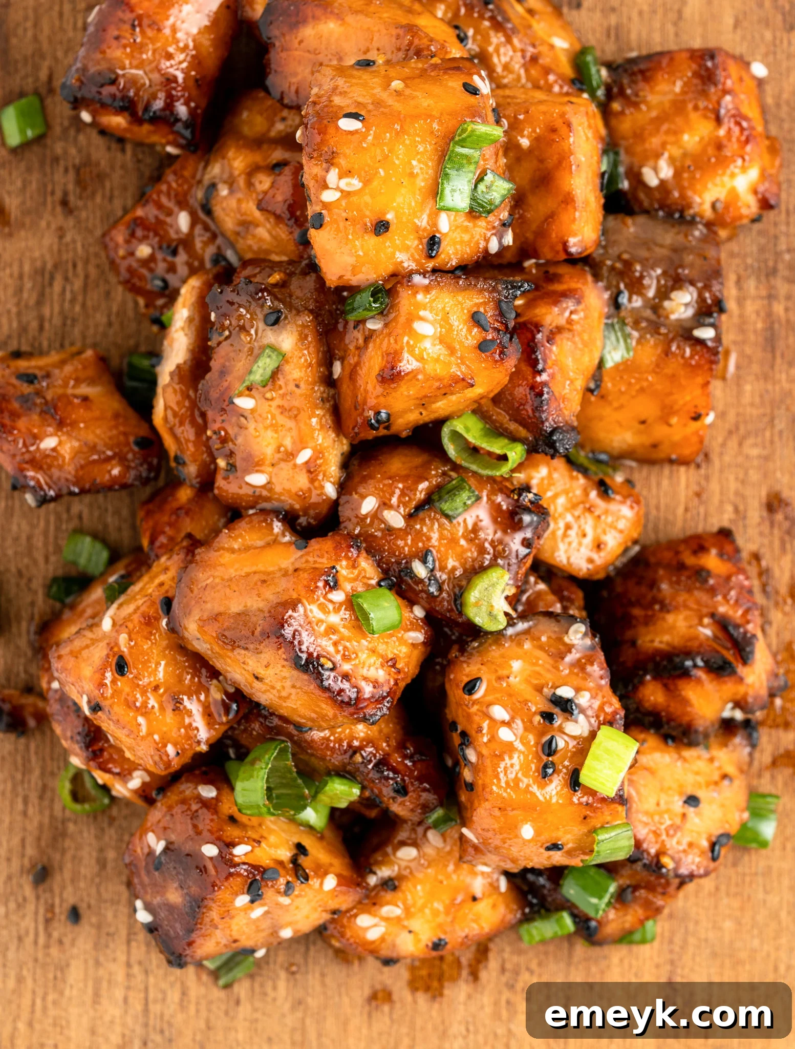 A pile of glazed, air fryer salmon bites topped with sesame seeds and sliced green onions on a wooden surface. The salmon has a caramelized appearance, indicating its well-cooked and flavorful.