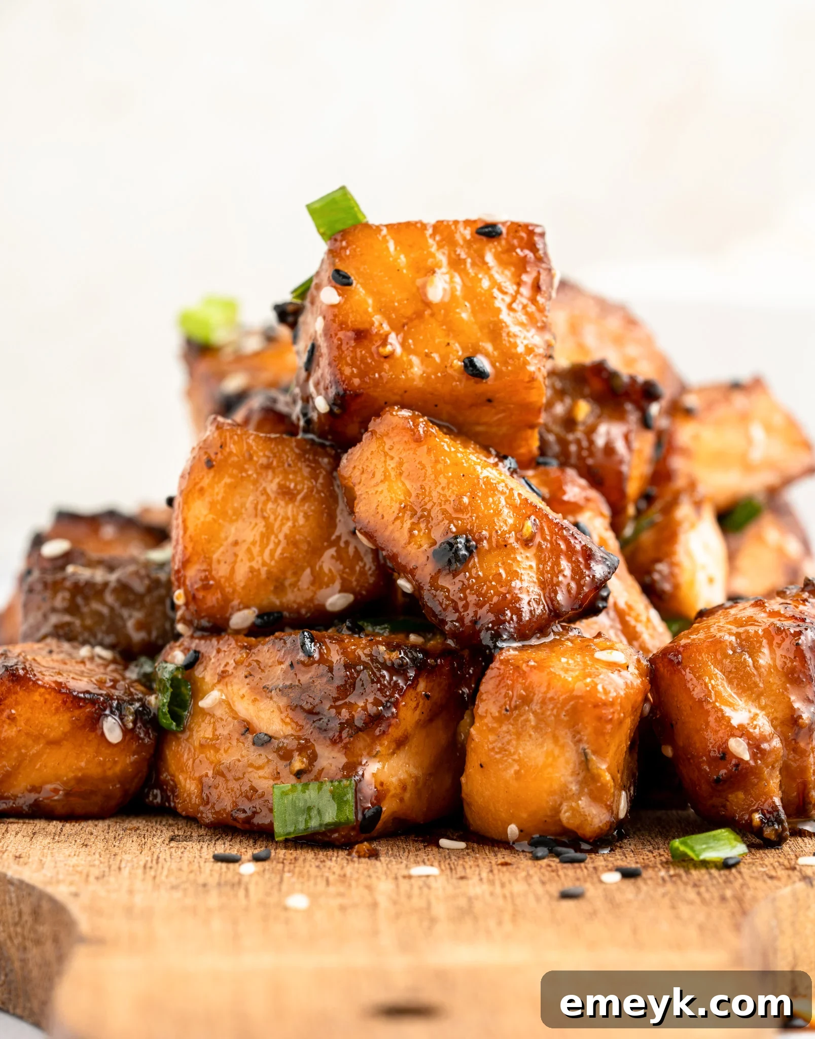 A stack of air fryer salmon bites garnished with sesame seeds and green onions on a wooden board. The pieces are golden-brown, highlighting their caramelized surface.